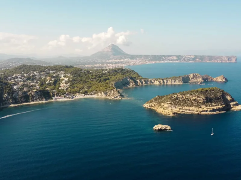 Panorama aérien de la côte de Jávea, révélant des collines verdoyantes avec des résidences, une plage de sable et une île rocheuse dans la mer bleue. Un voilier et le sillage d'un bateau sont visibles, avec le Montgó en arrière-plan. Panorama aérien de la côte de Jávea, révélant des collines verdoyantes avec des résidences, une plage de sable et une île rocheuse dans la mer bleue. Un voilier et le sillage d'un bateau sont visibles, avec le Montgó en arrière-plan.