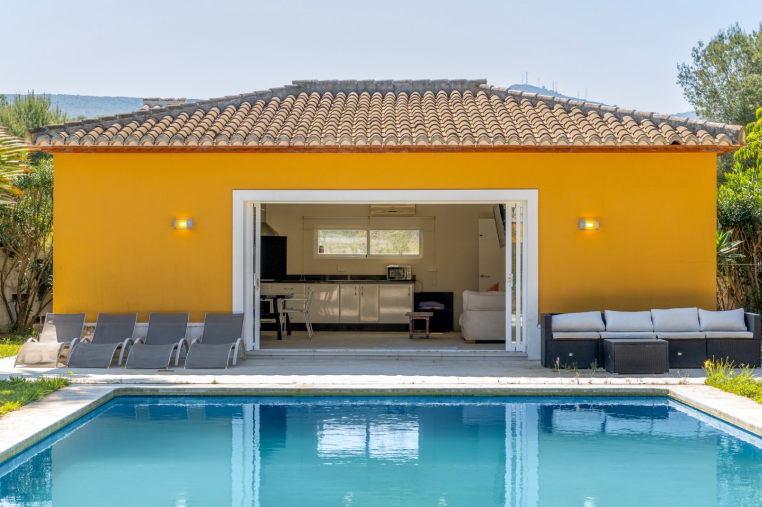 Annexe de piscine comprenant une cuisine d'été et un salon, ouverte sur une large terrasse ensoleillée et un bassin rectangulaire bordé de transats.