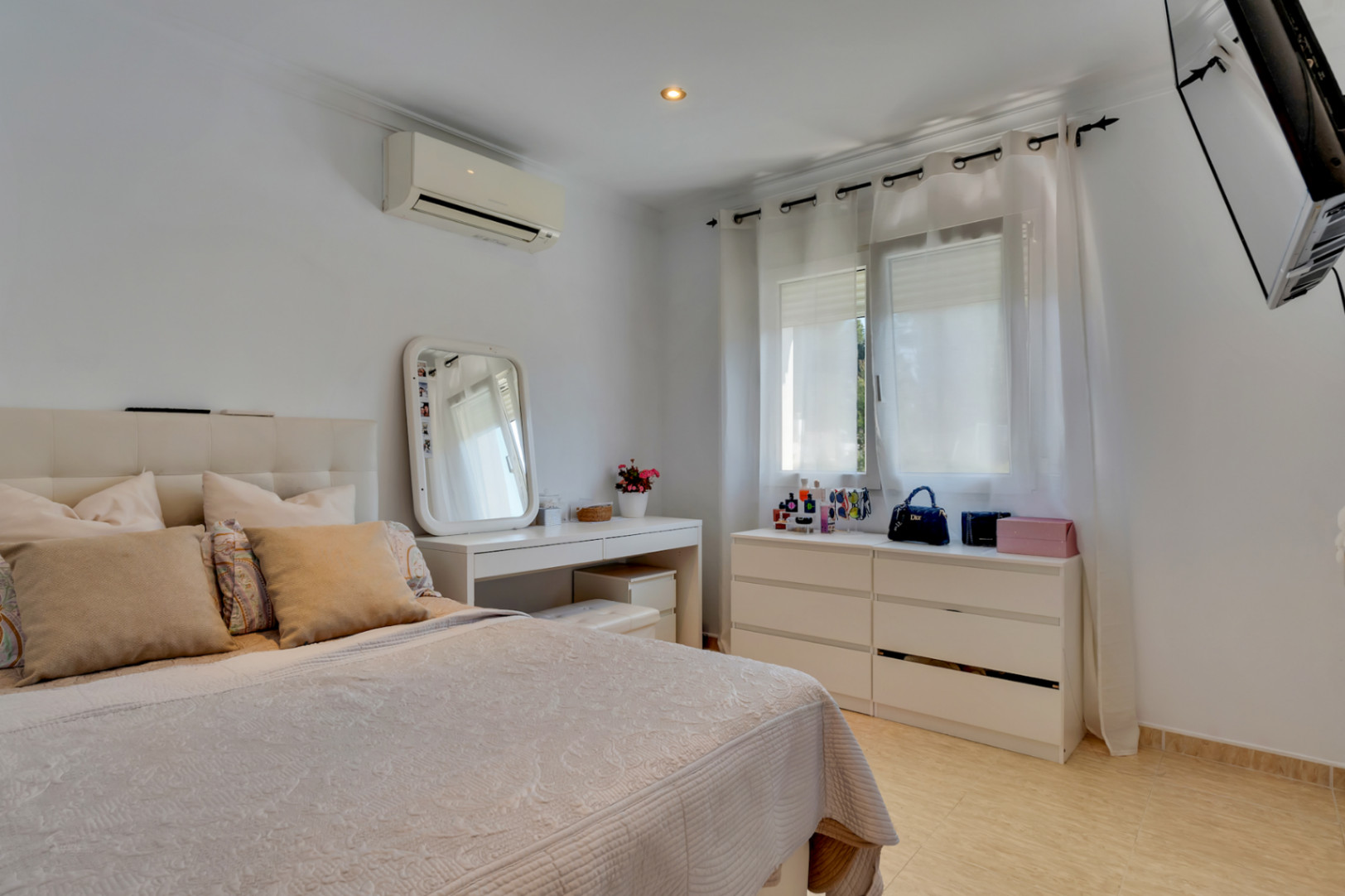 Chambre lumineuse avec mobilier blanc, coiffeuse, climatisation réversible et sol en grès. Une fenêtre avec rideaux voilages apporte une clarté naturelle à la pièce.