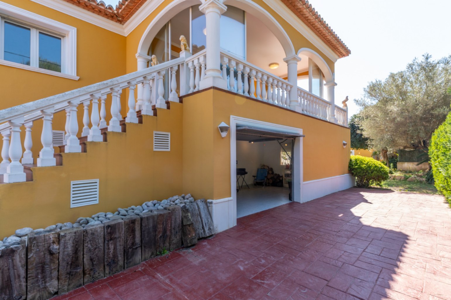 Façade de style méditerranéen avec escalier extérieur et balustrade blanche menant à une terrasse voûtée surplombant un garage intégré et une cour pavée.
