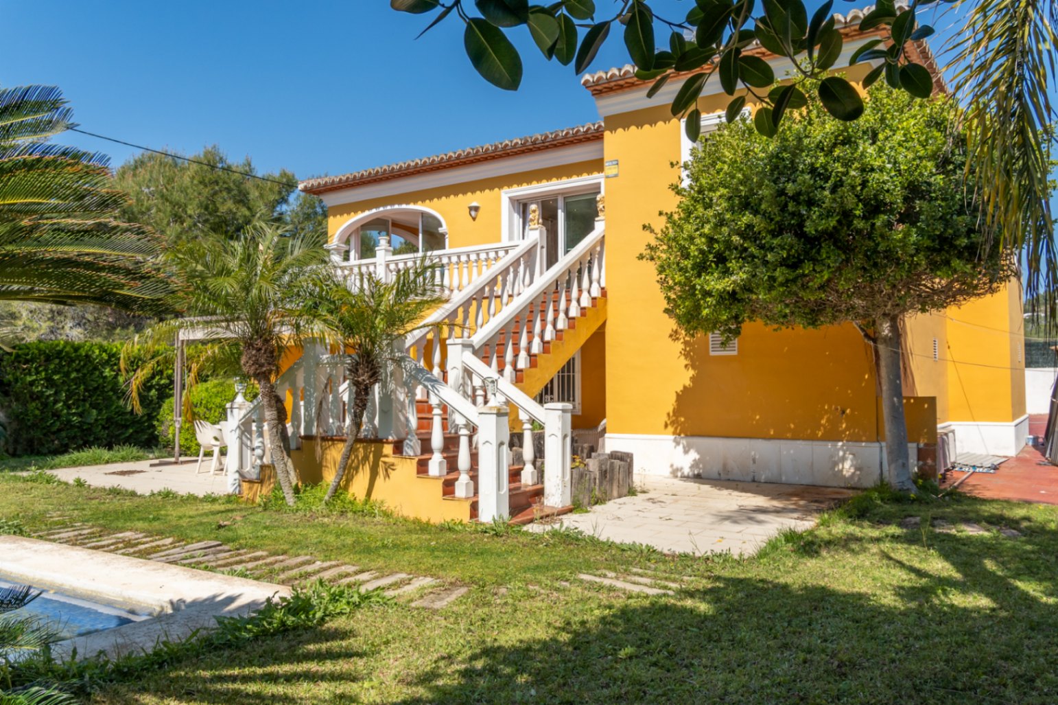 Façade jaune avec escalier extérieur à balustrade blanche menant à une terrasse couverte. Jardin méditerranéen avec palmiers et bord de piscine visible.
