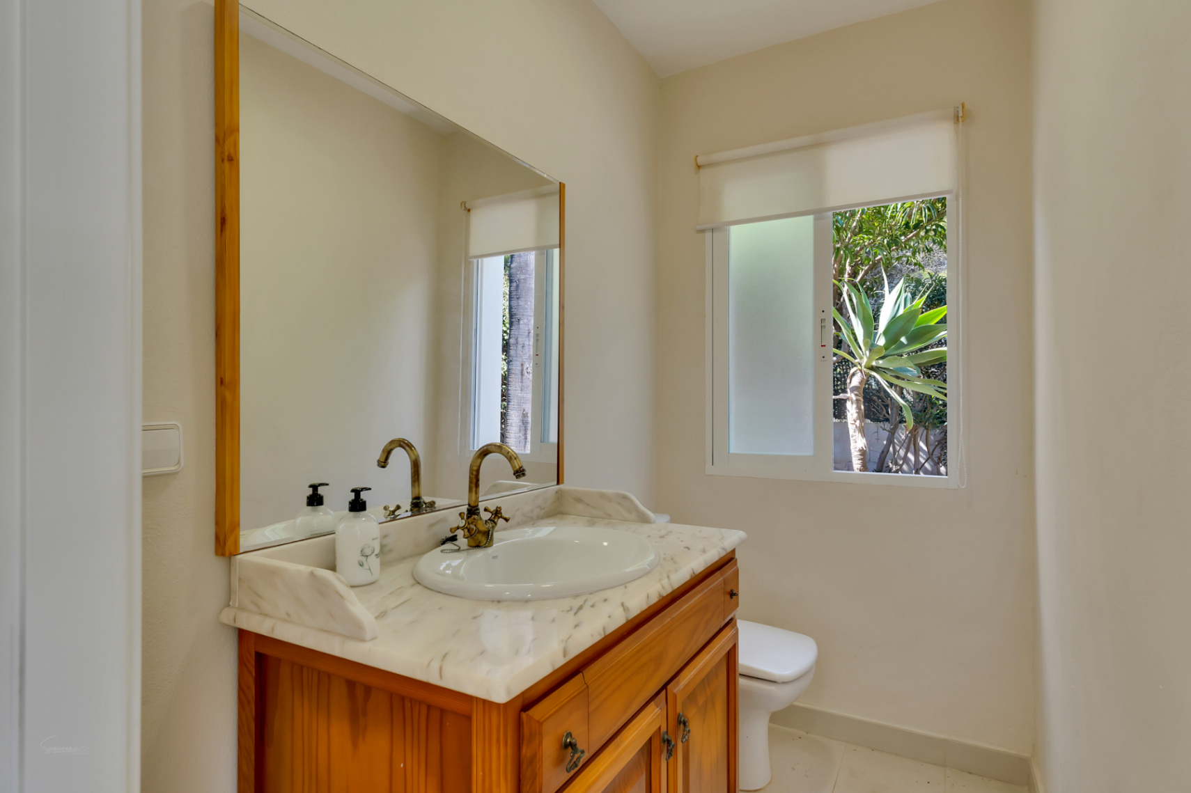 Salle de bain avec meuble en bois, plan de travail en marbre et robinetterie classique. Fenêtre offrant une lumière naturelle et vue sur la végétation.