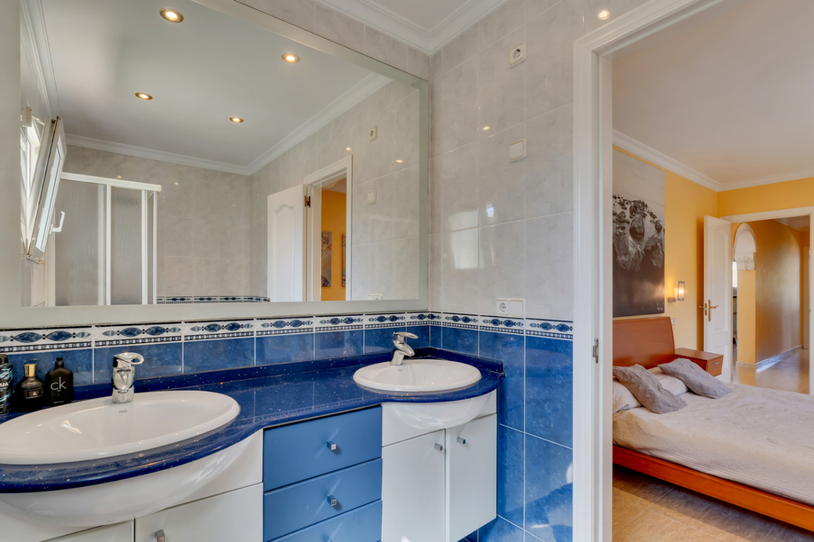 Salle de bains avec double vasque sur plan de travail bleu, carrelage décoratif et grand miroir. Accès direct à une chambre lumineuse aux tons chauds.