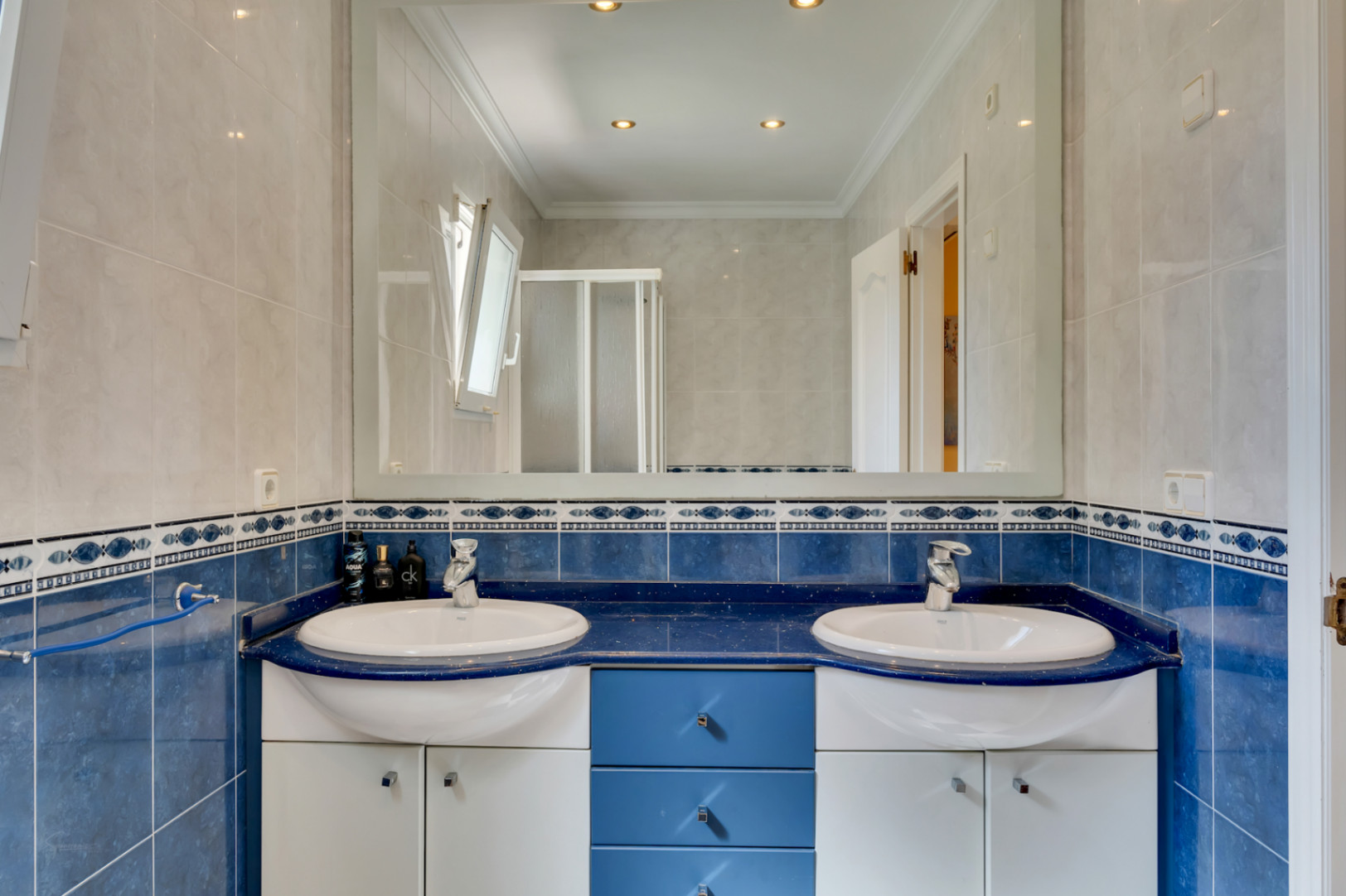 Salle de bains avec double vasque sur plan de travail bleu, grand miroir mural, éclairage encastré et carrelage mural bicolore à frise décorative.