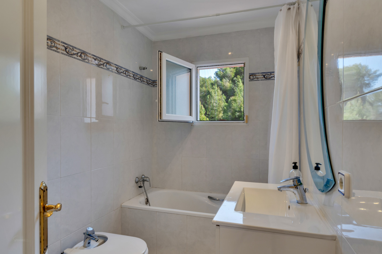 Salle de bains fonctionnelle avec baignoire encastrée, bidet et meuble vasque. Une fenêtre offre une vue sur la nature, soulignée par un carrelage avec liseré décoratif.