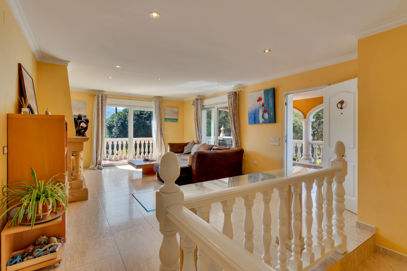 Salon spacieux avec carrelage clair et murs ocres, s'ouvrant sur une terrasse par de grandes baies vitrées. Une balustrade blanche délimite cet espace de vie convivial.