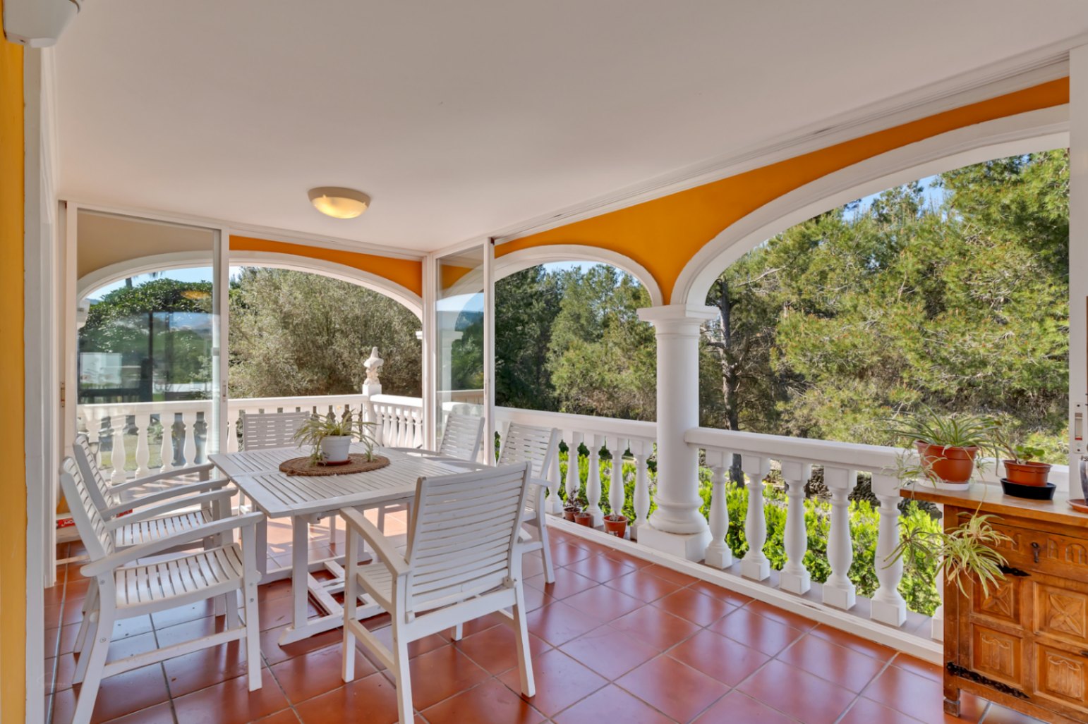 Terrasse couverte avec arches, balustrade blanche et sol en terre cuite. Cet espace comprend un coin repas en bois blanc offrant une vue dégagée sur la pinède environnante.