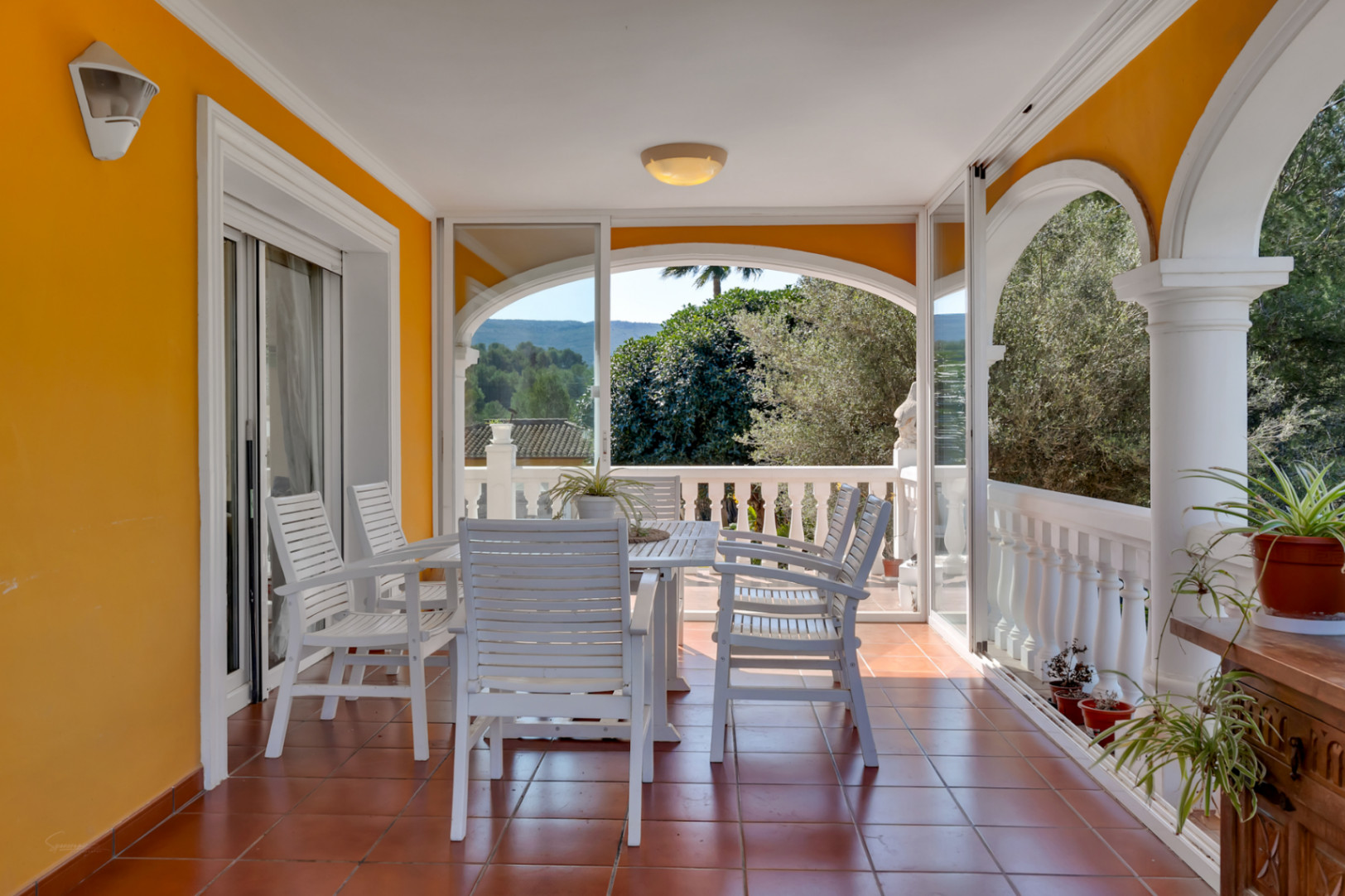 Terrasse couverte avec coin repas, sol en terre cuite et baies vitrées offrant une vue panoramique sur la végétation environnante et les collines.
