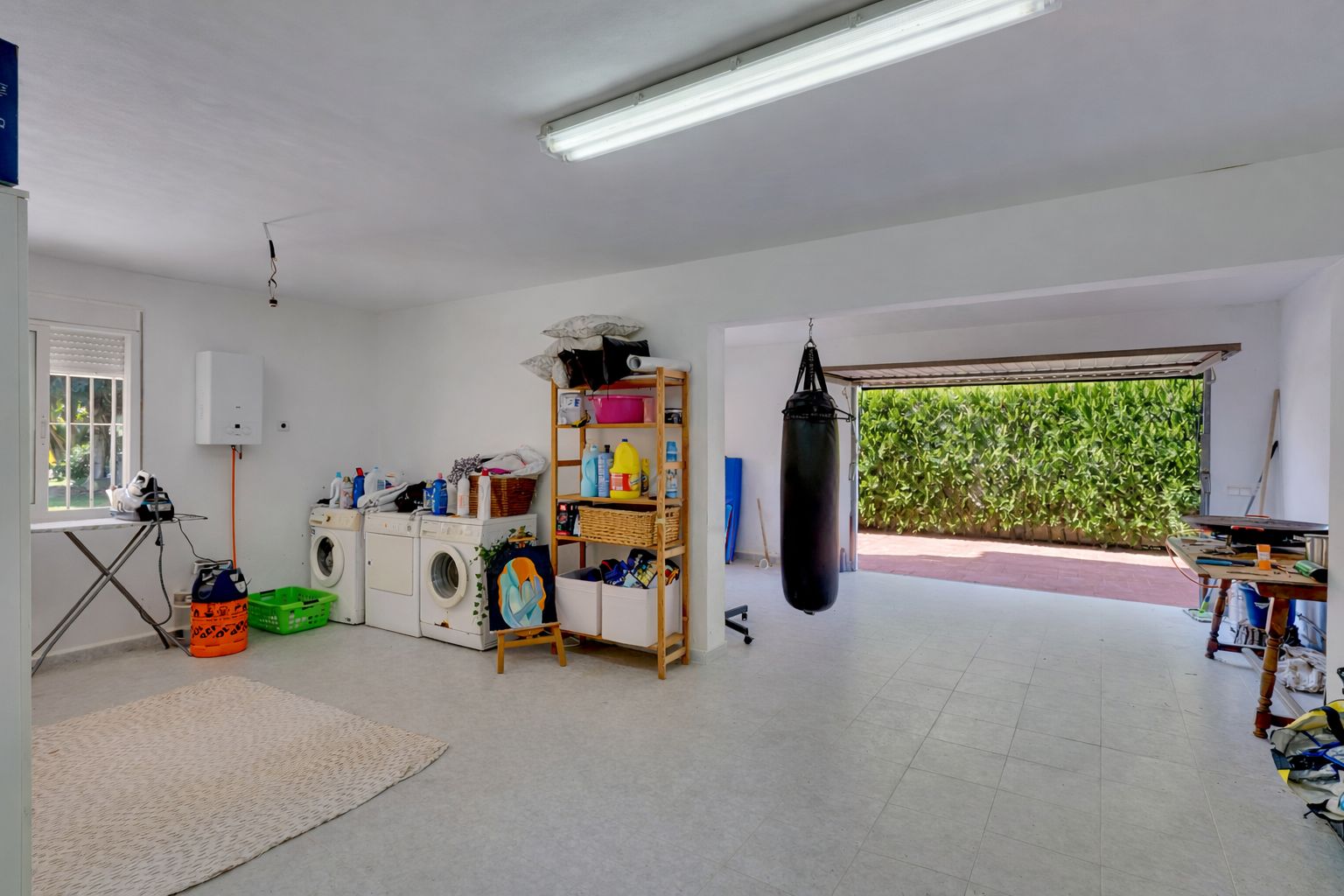 Vaste espace polyvalent comprenant une buanderie équipée, des rangements et un coin fitness avec sac de frappe, ouvrant sur l'extérieur par la porte de garage.