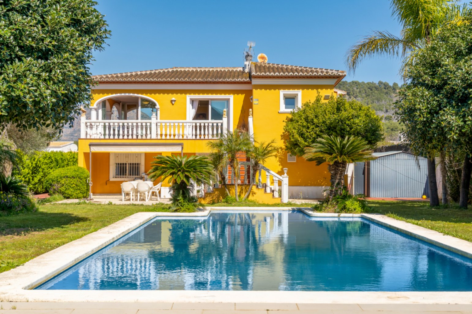 Villa méditerranéenne avec façade jaune, grande piscine rectangulaire, terrasse à balustrade blanche et jardin paysager avec palmiers et pelouse.