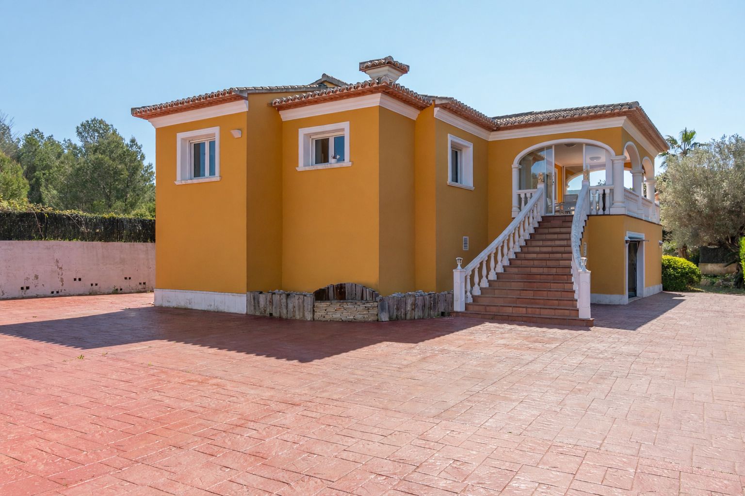 Vue extérieure de la villa ocre avec son grand escalier à balustrade blanche menant à une terrasse couverte sur une vaste cour en béton imprimé.