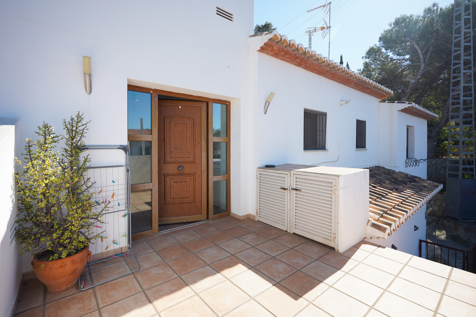 Accès indépendant au logement supérieur avec porte en bois sculpté, terrasse en grès rustique et rangements extérieurs intégrés sur façade blanche.