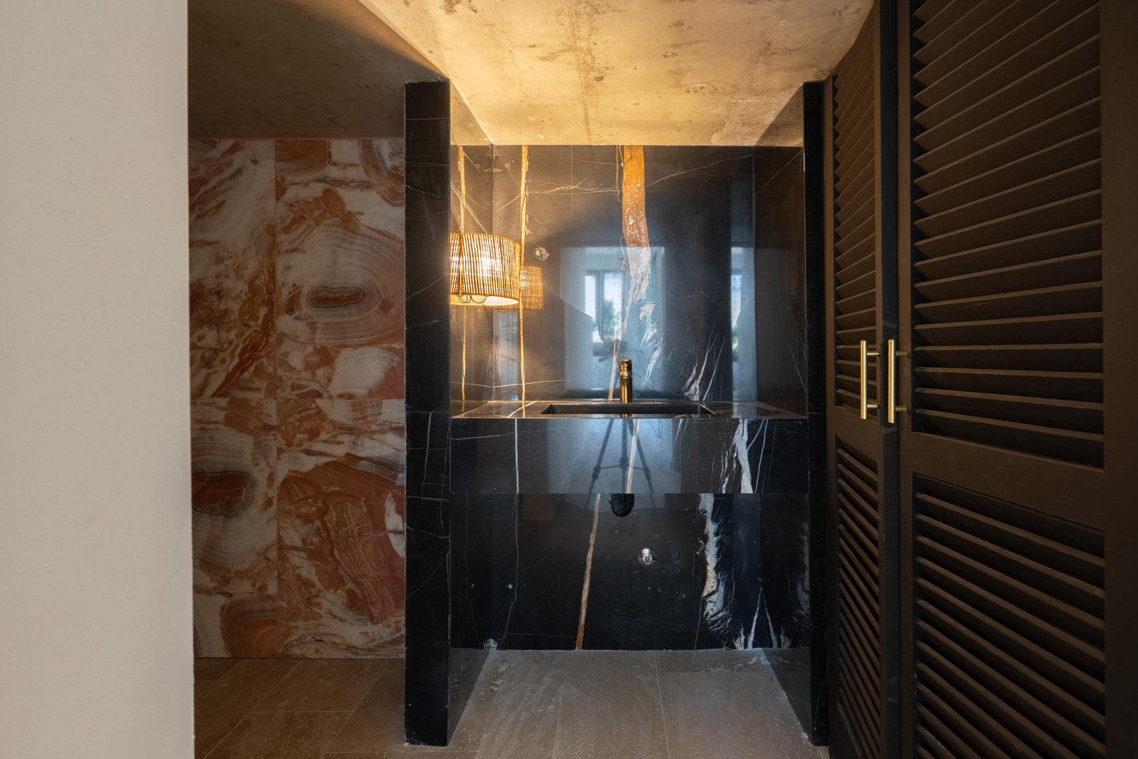 Cabinet de toilette design avec lavabo intégré en marbre noir veiné, robinetterie dorée, murs en pierre naturelle et portes à persiennes sombres aux poignées dorées.