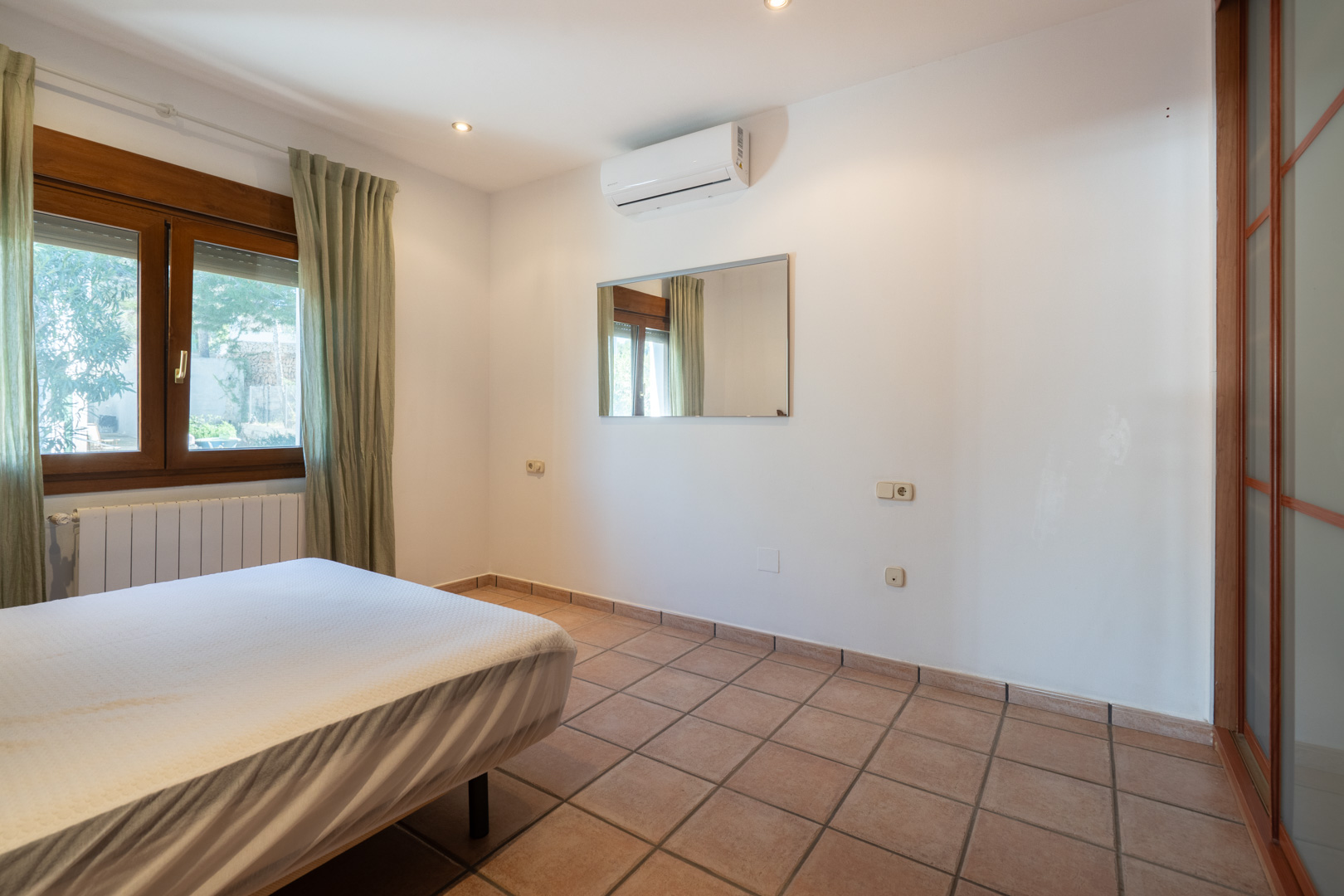 Chambre avec sol en carrelage, climatisation split, fenêtre en bois et placard intégré doté de portes coulissantes translucides.