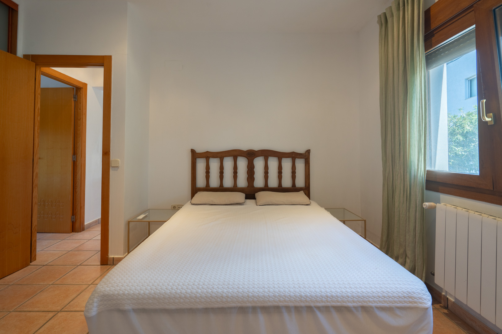 Chambre avec sol en terre cuite, tête de lit en bois sculpté et fenêtre à cadre en bois. La pièce dispose de tables de chevet en verre et d'un radiateur blanc.