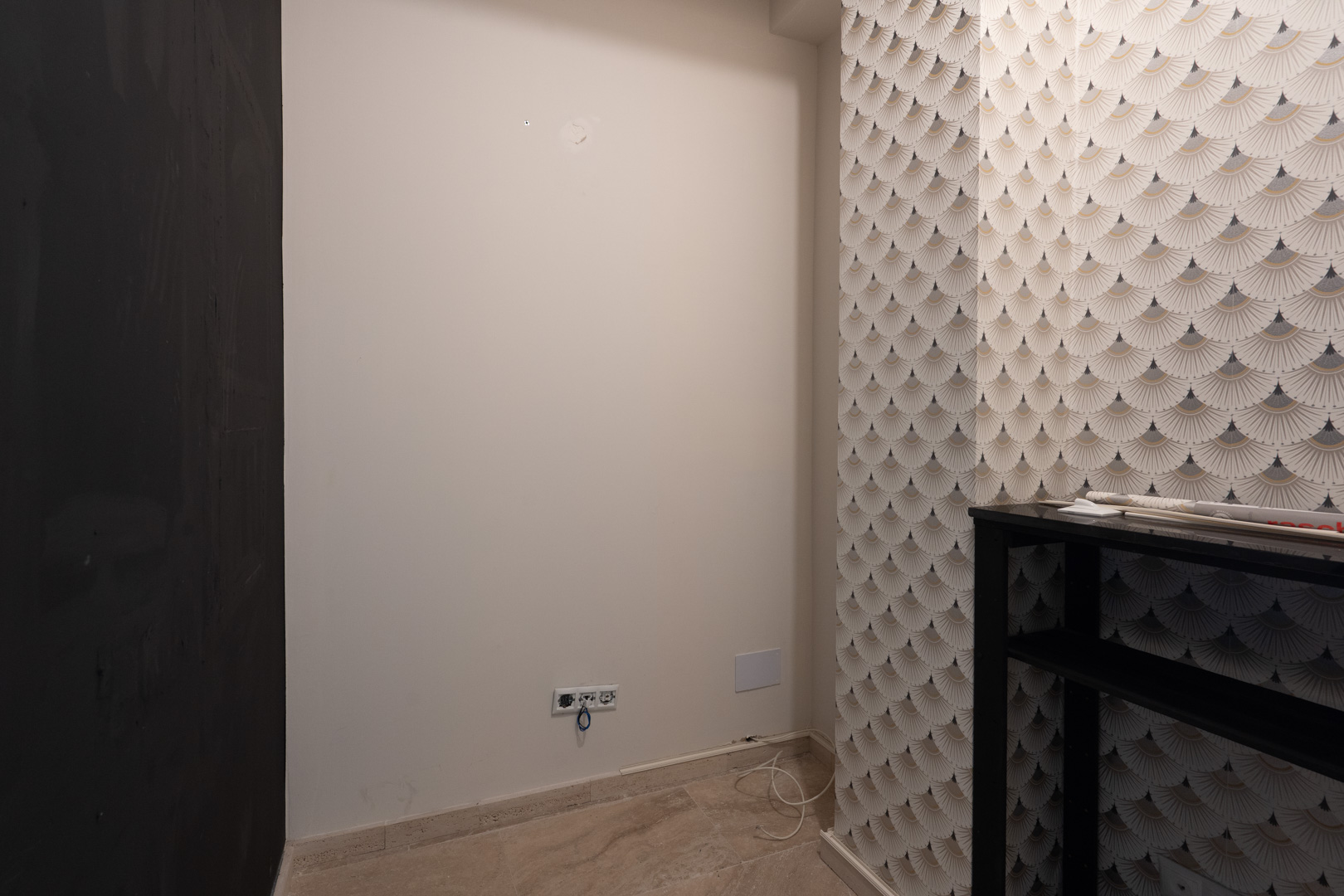 Coin intérieur avec papier peint à motifs géométriques, murs blancs neutres équipés de prises électriques et revêtement de sol en pierre claire.
