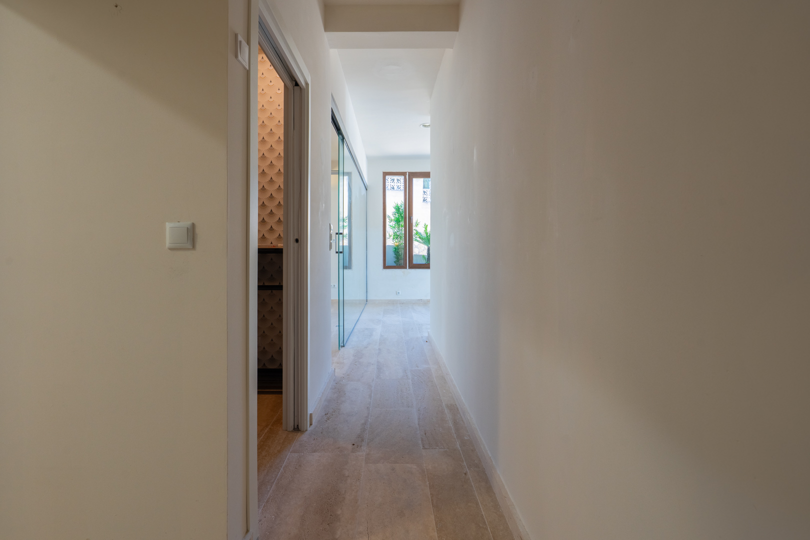 Couloir de distribution avec sol en pierre, murs neutres et accès à des pièces dotées de papier peint design et de cloisons vitrées.