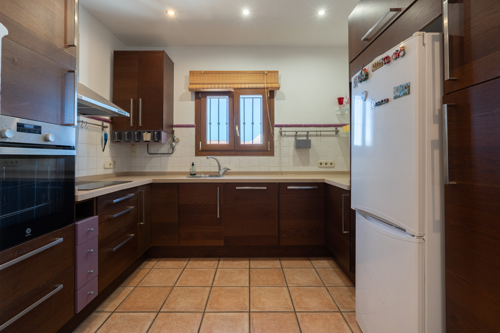 Cuisine fonctionnelle avec des armoires en bois sombre, un plan de travail beige et un sol en grès. Équipée d'un four encastré et d'un éclairage encastré au plafond.
