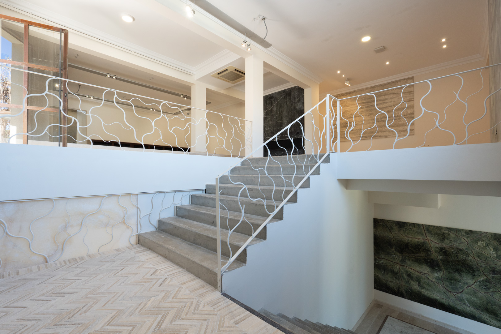 Escalier architectural avec garde-corps blanc aux motifs organiques, reliant les niveaux commerciaux spacieux dotés de sols en chevrons et de panneaux muraux en pierre.