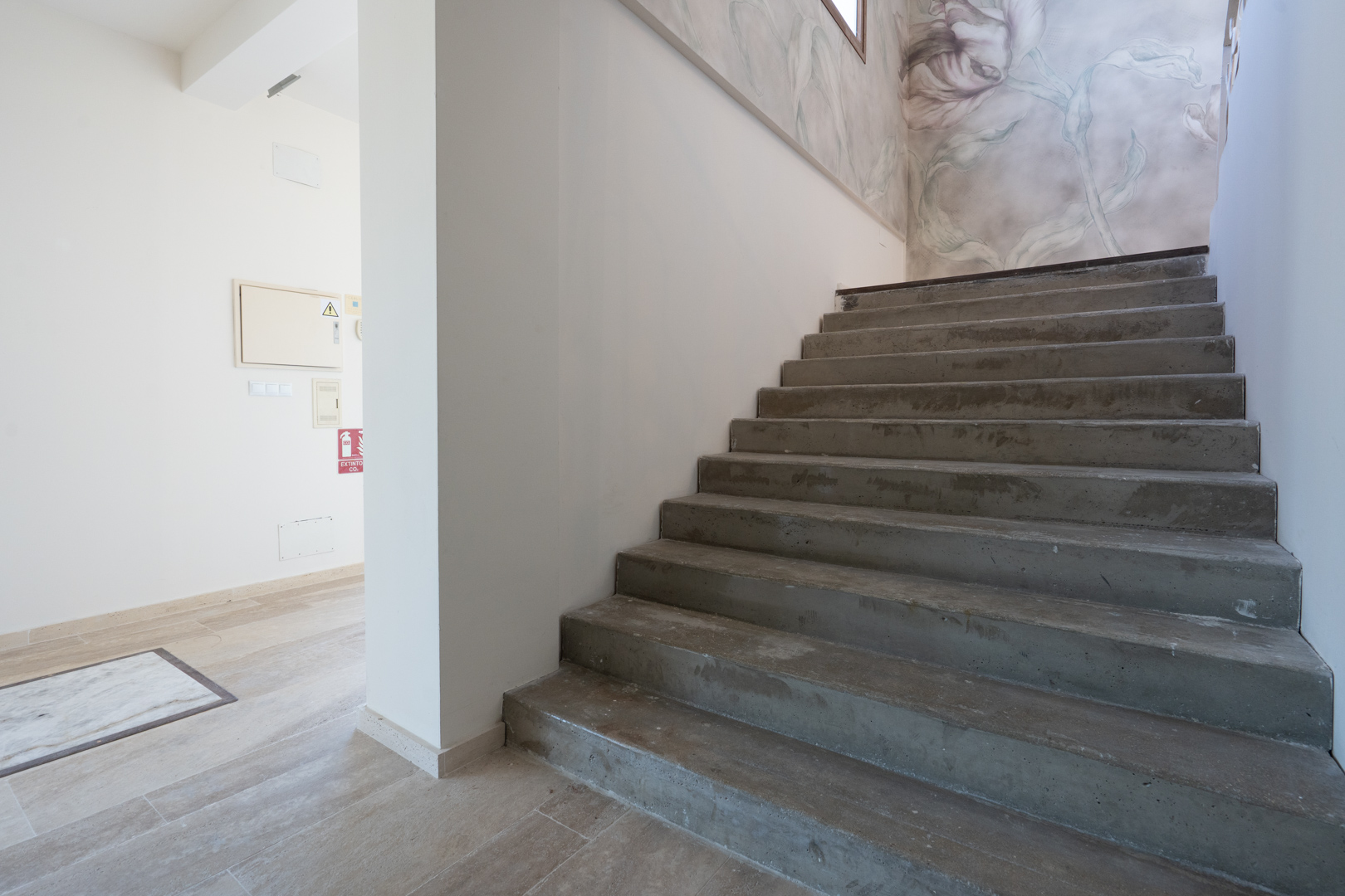 Escalier intérieur en béton avec une fresque murale florale artistique, situé à côté d'un hall d'entrée aux sols en pierre claire.