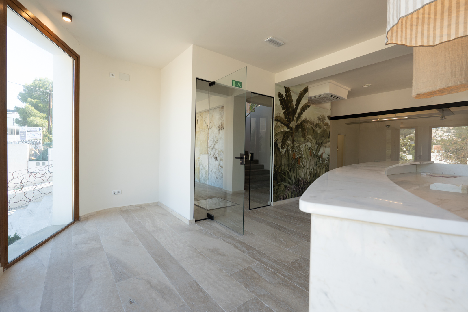 Espace de réception commercial avec comptoir courbé en marbre blanc, sol en grès cérame, portes vitrées et fresque murale aux motifs tropicaux.