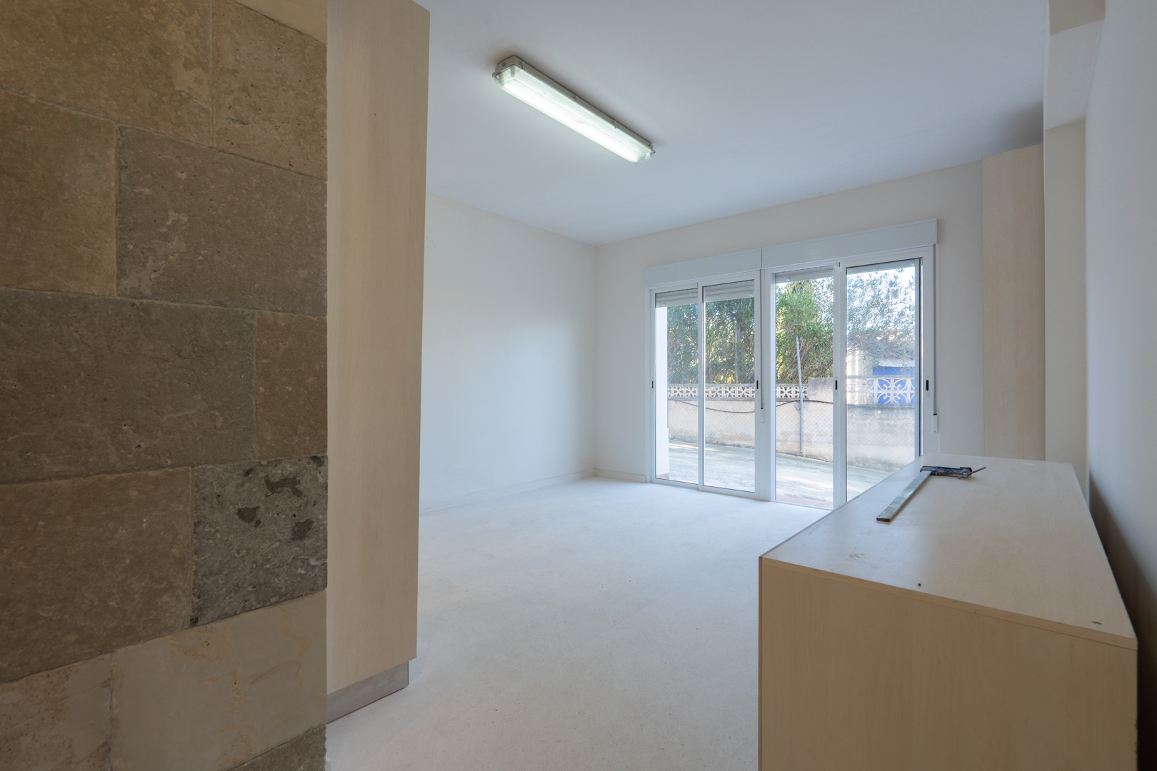 Espace polyvalent au rez-de-chaussée avec mur en pierre naturelle, éclairage fonctionnel et accès extérieur par de larges baies vitrées coulissantes.