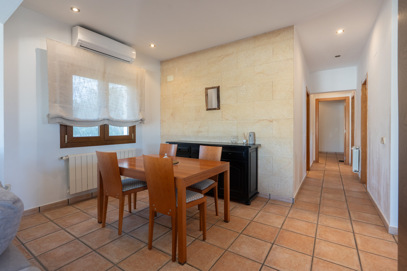 Espace salle à manger avec mur en pierre naturelle, sol en terre cuite, climatisation et couloir desservant les chambres de l'appartement.