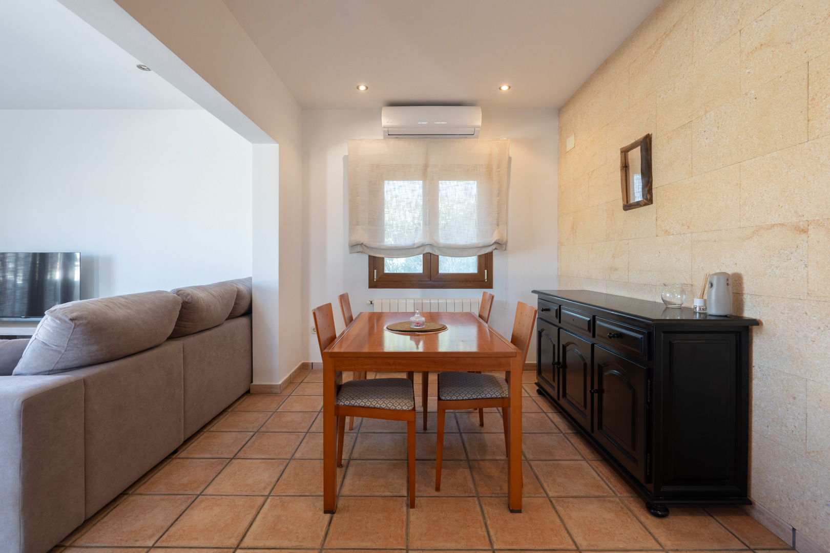 Espace salle à manger avec mur en pierre naturelle, sol en terre cuite, table en bois pour quatre et buffet noir. Équipé de la climatisation et du chauffage.