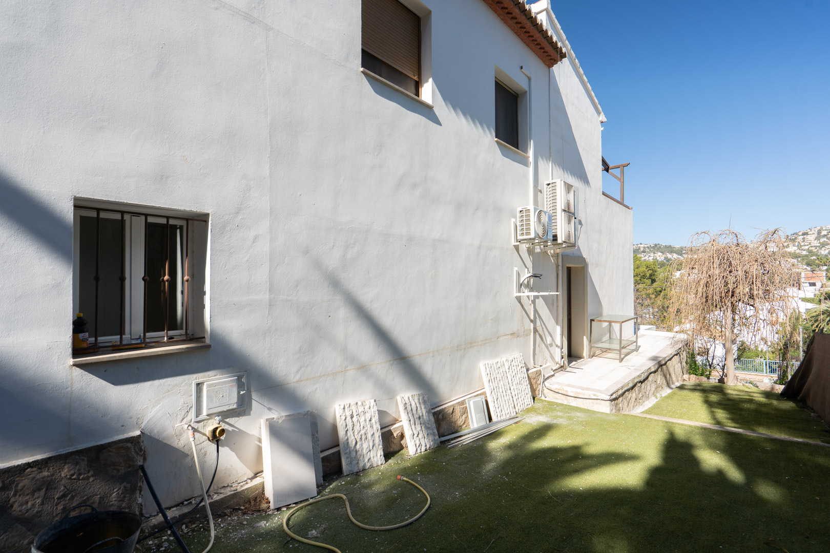 Façade latérale blanche avec soubassement en pierre naturelle, unités de climatisation extérieures et espace jardin avec gazon synthétique offrant une vue dégagée.