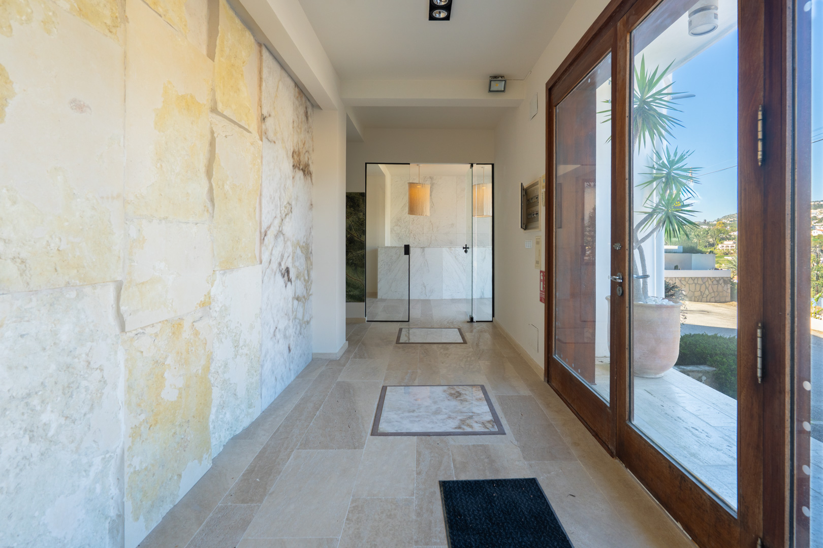 Hall d'entrée avec murs en pierre naturelle, sol en travertin avec inserts en marbre et portes vitrées menant à l'espace de réception.