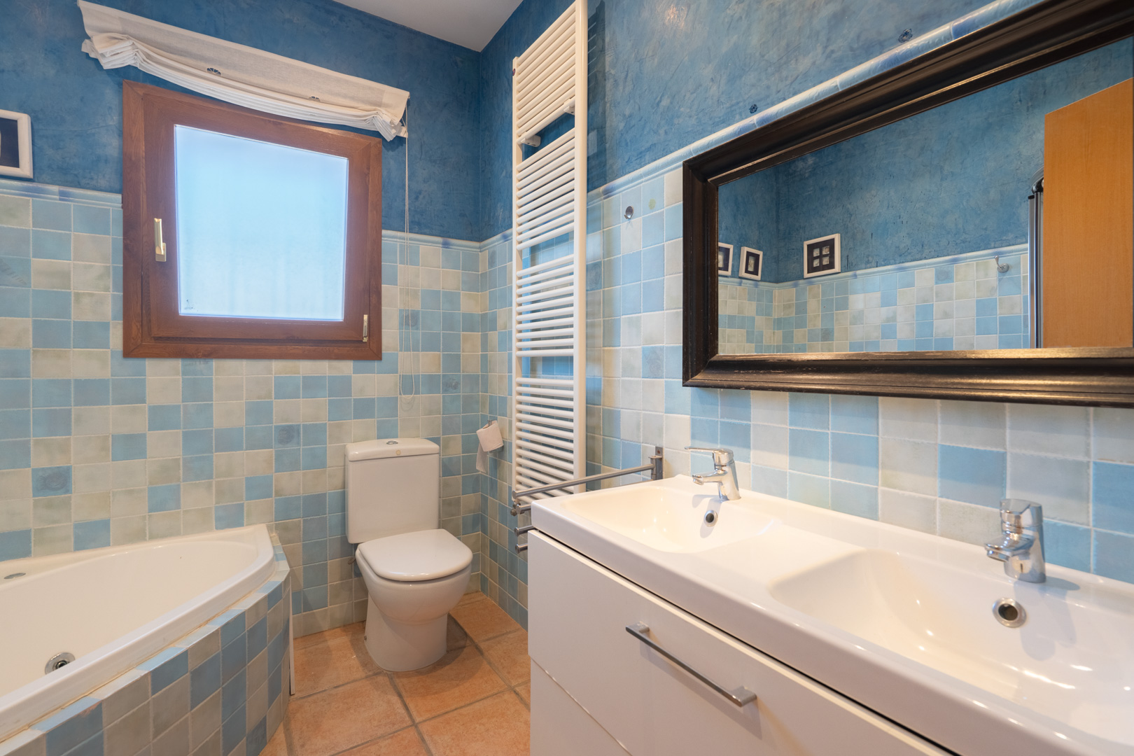 Salle de bain avec carrelage en damier bleu, double vasque moderne, baignoire d'angle et radiateur sèche-serviettes blanc sur murs à effet texturé.