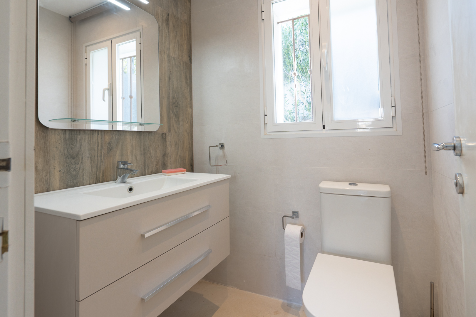 Salle de bain moderne avec meuble vasque suspendu, mur d'accent aspect bois, miroir avec étagère en verre et fenêtre en PVC apportant de la lumière naturelle.