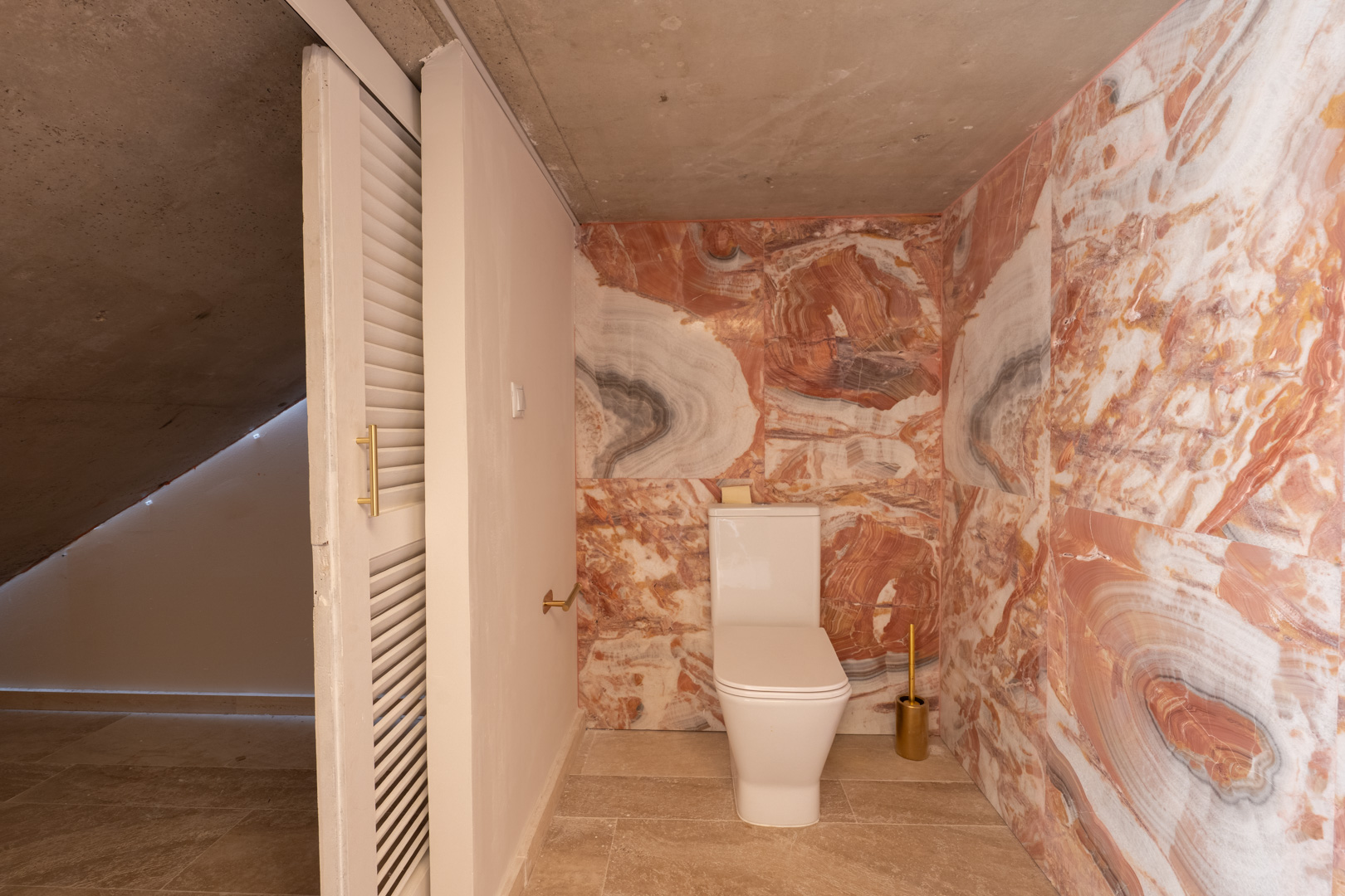 Toilettes invités avec murs en marbre veiné rouge, WC moderne et plafond en béton brut, le tout souligné par une porte coulissante à persiennes.