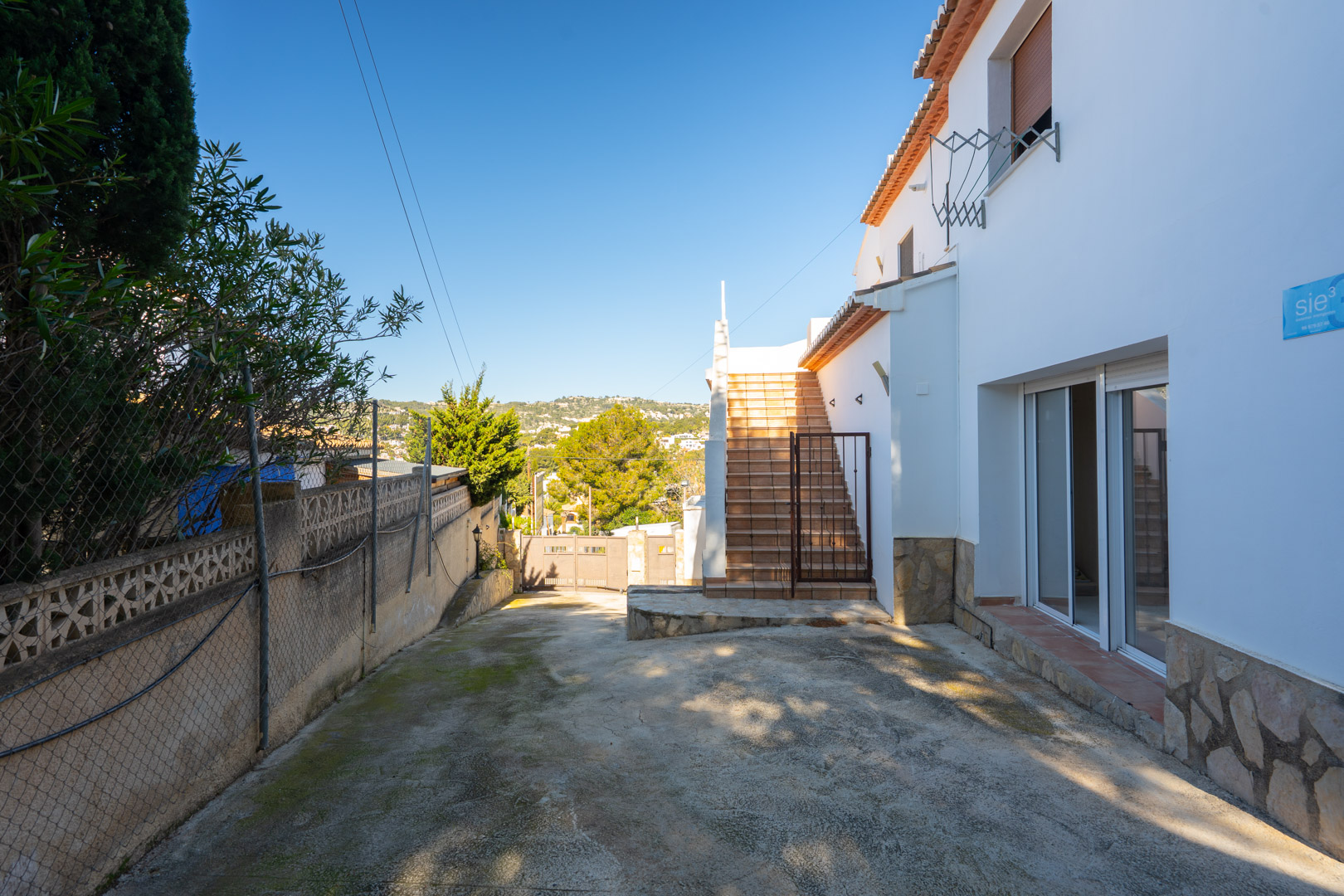 Vue extérieure montrant l'accès indépendant au premier étage par un escalier extérieur, une zone de stationnement privée et une façade blanche avec soubassement en pierre.