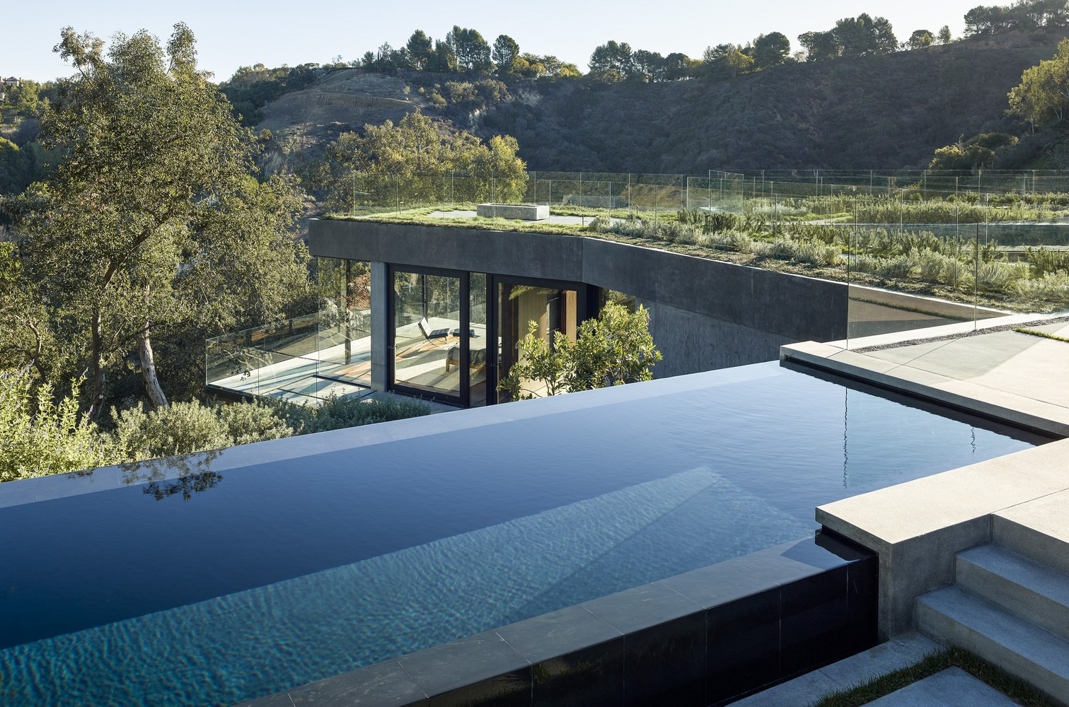 Extérieur d'une villa moderne avec piscine à débordement, toiture végétalisée et grandes baies vitrées offrant une vue dégagée sur le relief environnant.