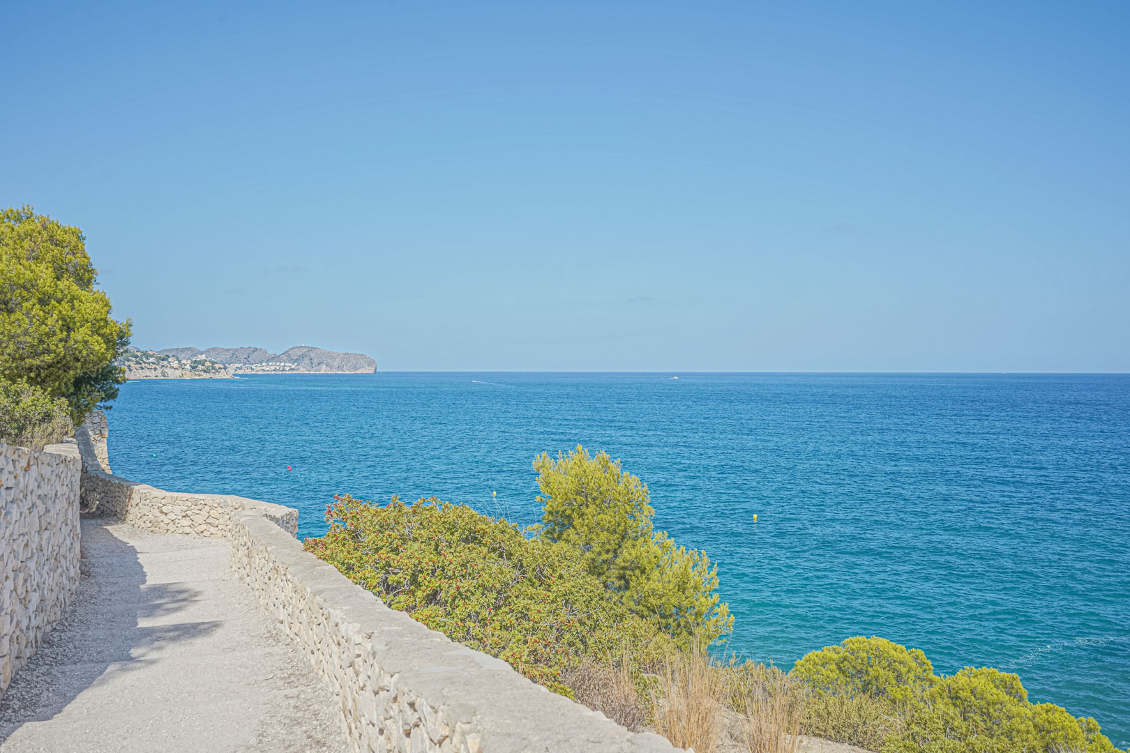 Sentier côtier avec des murs en pierre sèche offrant une vue panoramique sur la mer Méditerranée, bordé de pins et de végétation locale.