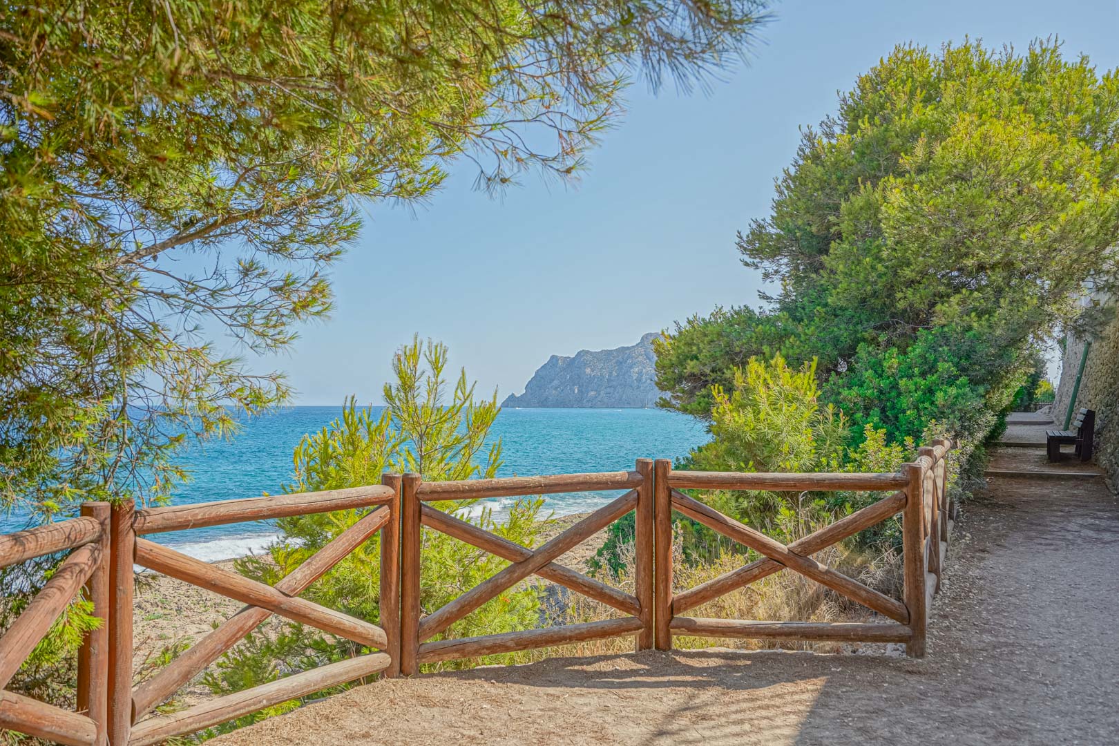 Sentier côtier avec garde-corps en bois offrant une vue panoramique sur la mer Méditerranée et le Peñón de Ifach, entouré de pins.