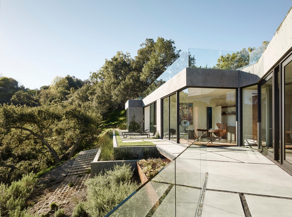 Villa moderne en béton avec baies vitrées toute hauteur et vaste terrasse aux garde-corps en verre, intégrée dans un paysage boisé.