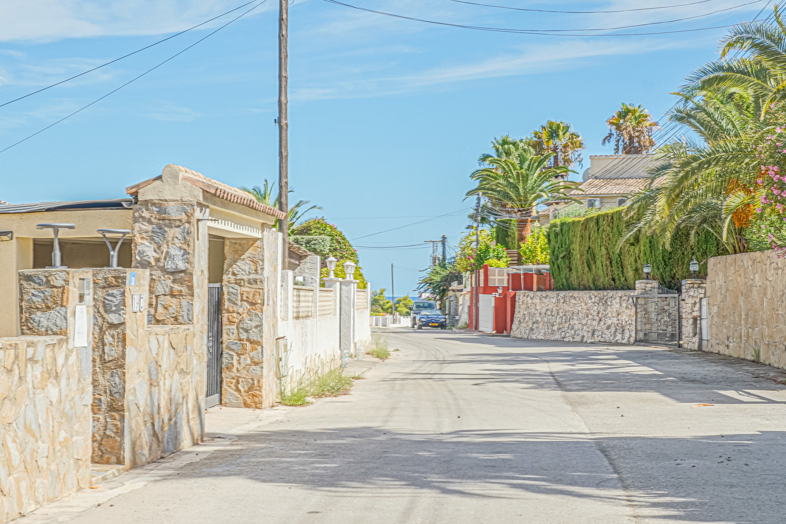 Vue de la rue résidentielle bordée de murs en pierre naturelle et de palmiers, avec un aperçu de la mer à l'horizon sous un ciel bleu.