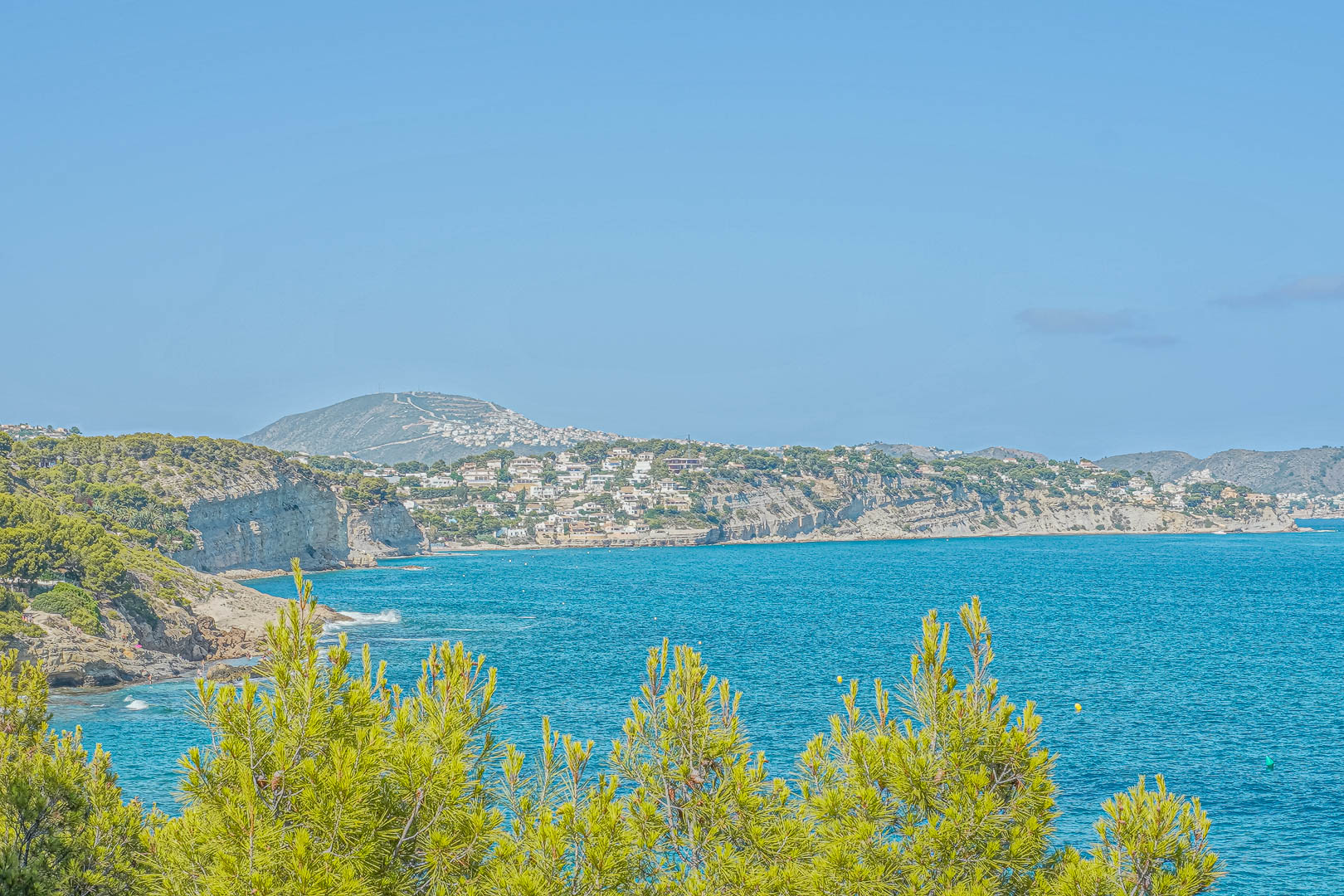 Vue panoramique sur la côte de Benissa avec des pins méditerranéens au premier plan et des falaises résidentielles surplombant les eaux turquoises de la mer.