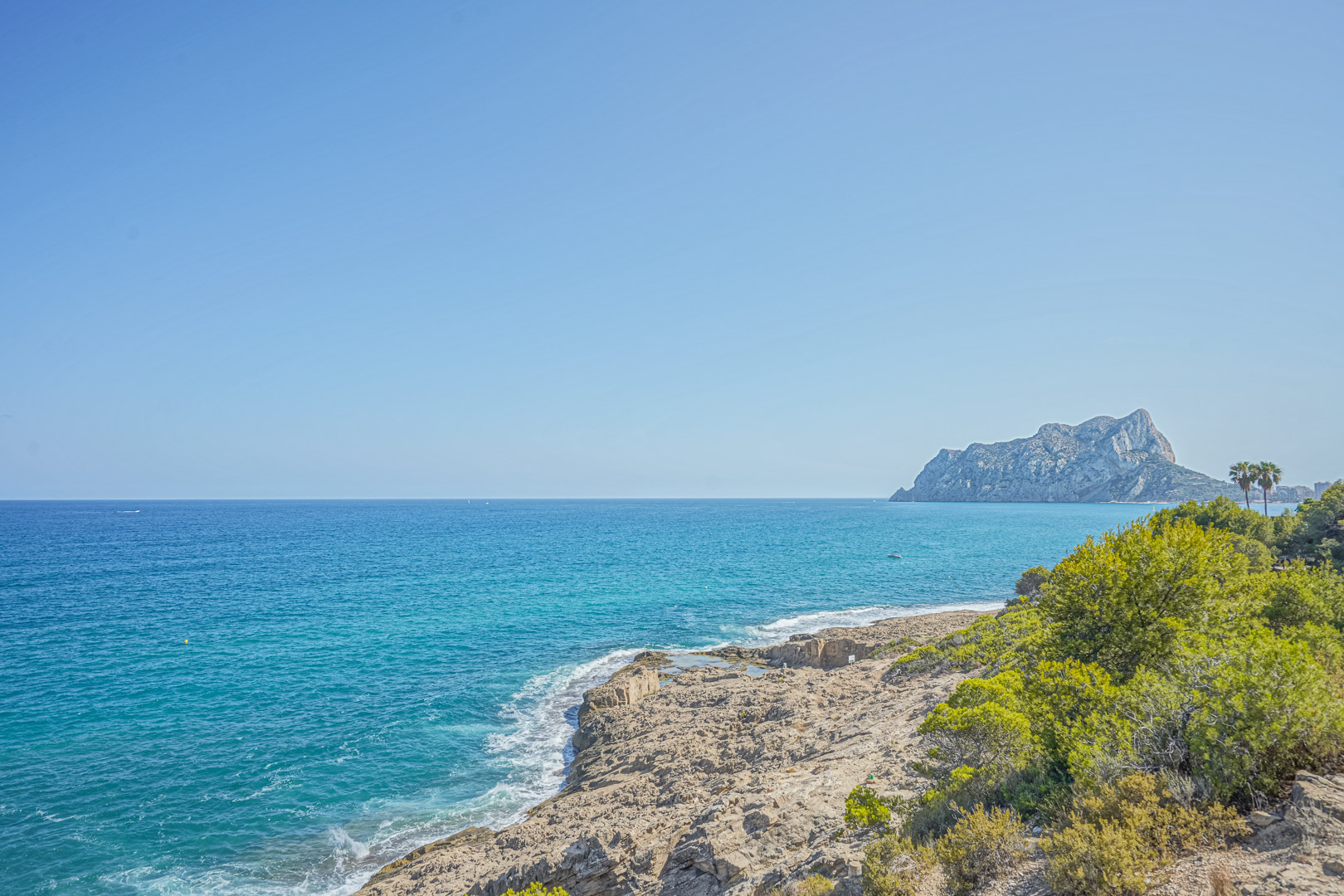 Vue panoramique sur la côte de Benissa avec le Peñón de Ifach à l'horizon, mettant en valeur les eaux turquoise et les falaises rocheuses bordées de pins.