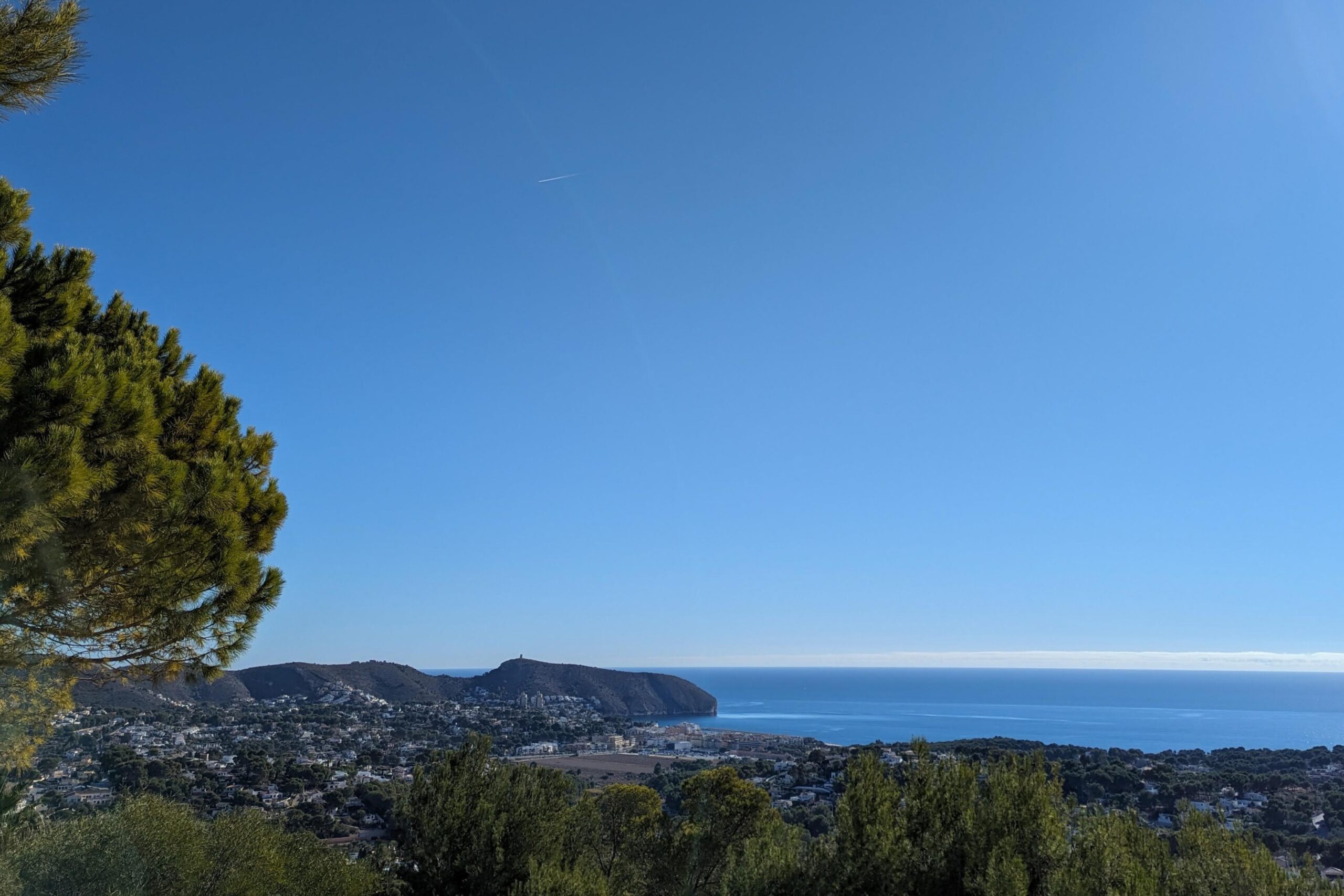 Vue panoramique surélevée sur la côte de Moraira et la mer Méditerranée, encadrée par des pins locaux sous un ciel bleu azur.