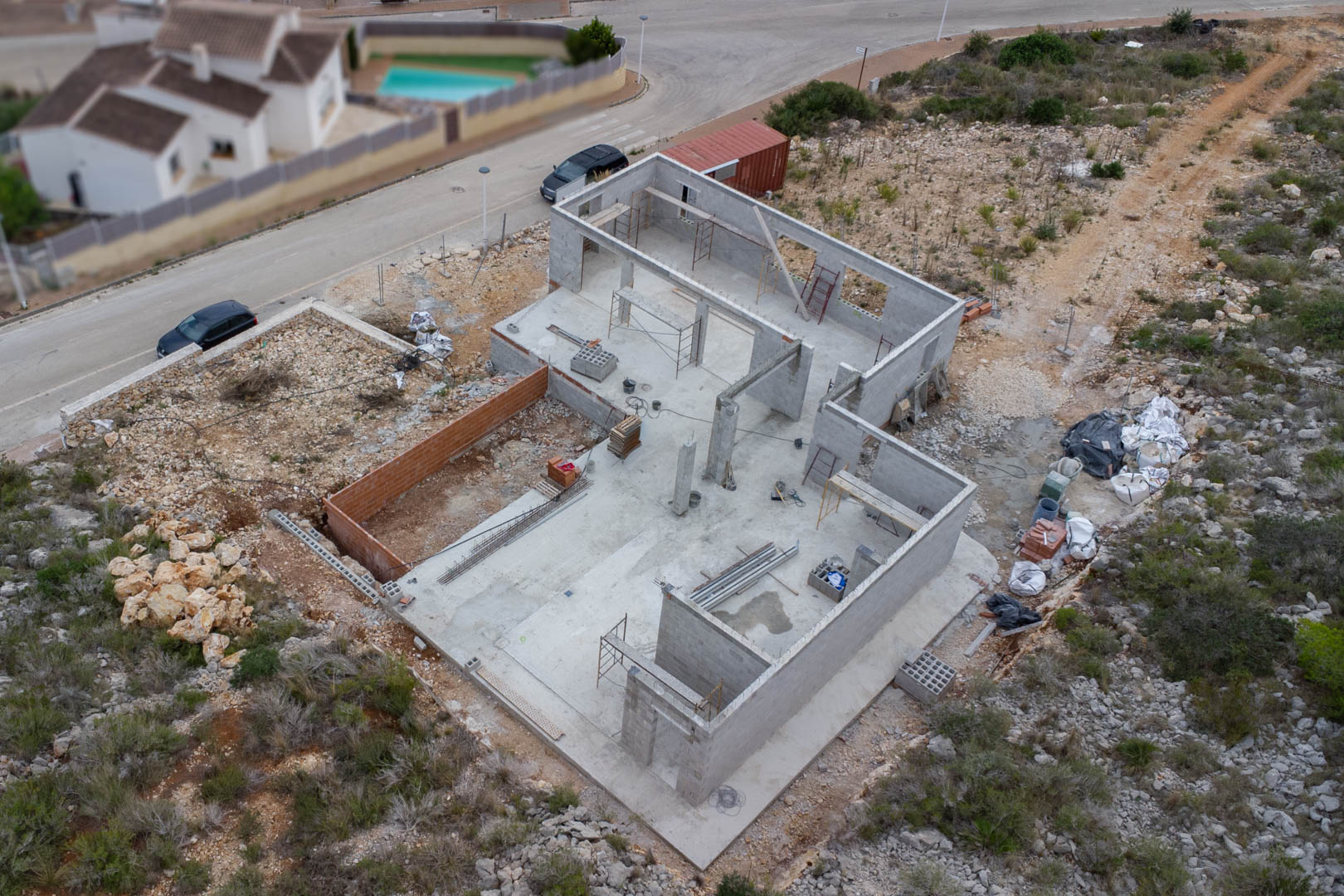 Vue aérienne des fondations en béton et de la structure des murs en parpaings de la villa en chantier, avec matériaux et échafaudages sur place.