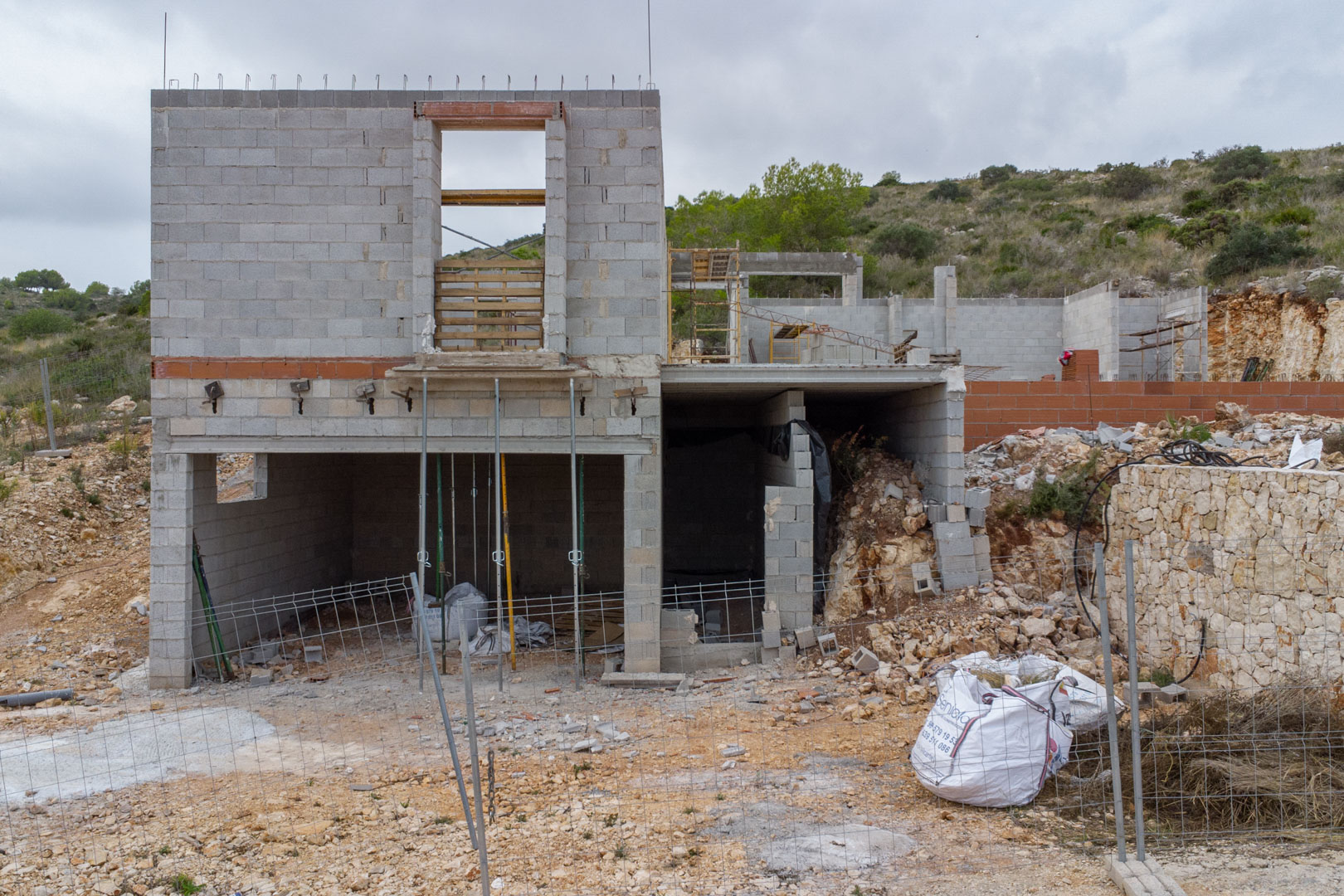 Vue extérieure de la structure en blocs de béton en construction, présentant plusieurs niveaux et un mur de soutènement en pierre naturelle sur une colline à Pedreguer.
