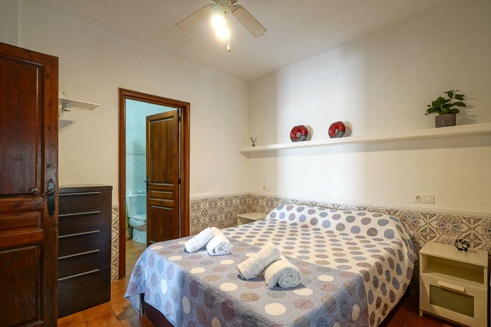 Chambre principale en suite avec soubassement en carreaux décoratifs, ventilateur de plafond et accès direct à la salle de bain attenante par une porte en bois.