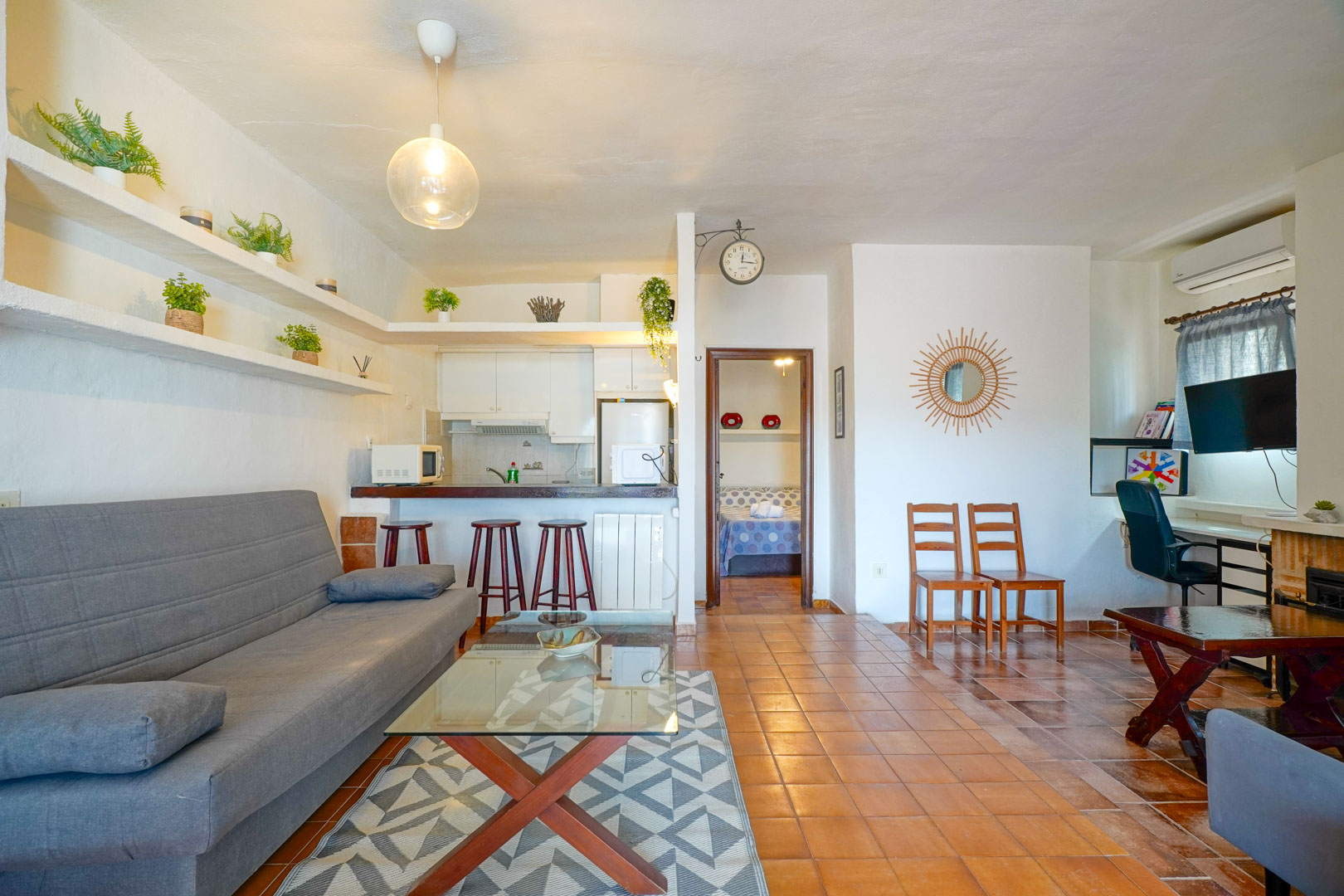 Espace de vie avec cuisine semi-ouverte, sol en terre cuite et étagères murales intégrées. La pièce comprend un coin salon, un bureau et une ouverture vers la suite principale.