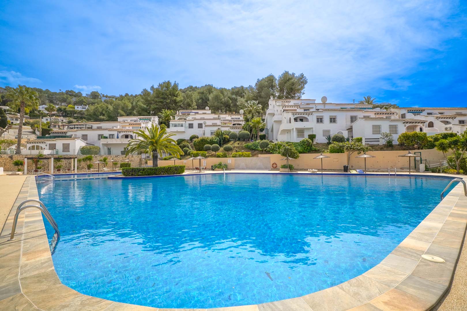 Grande piscine communautaire aux formes arrondies, entourée d'un solarium avec parasols en paille et d'une végétation luxuriante au pied des résidences de style méditerranéen.