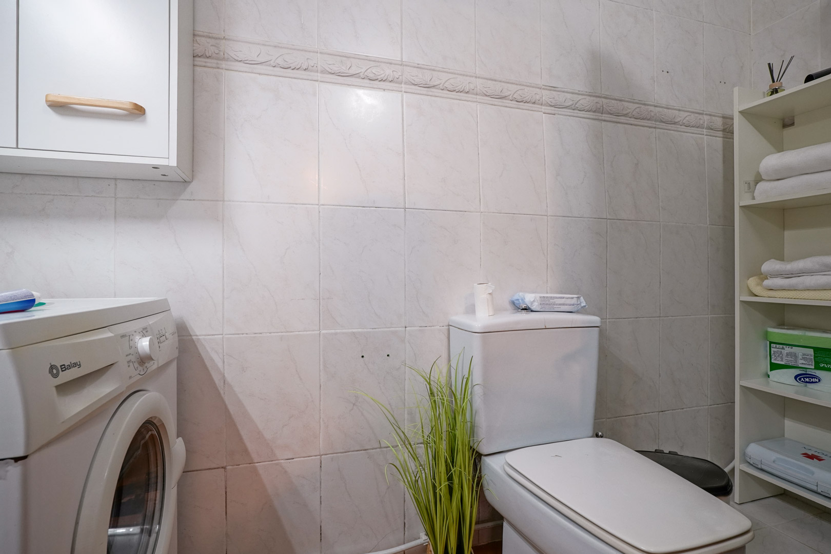 Salle de bain fonctionnelle avec coin buanderie, équipée d'un lave-linge Balay, d'étagères de rangement et de murs carrelés avec frise.