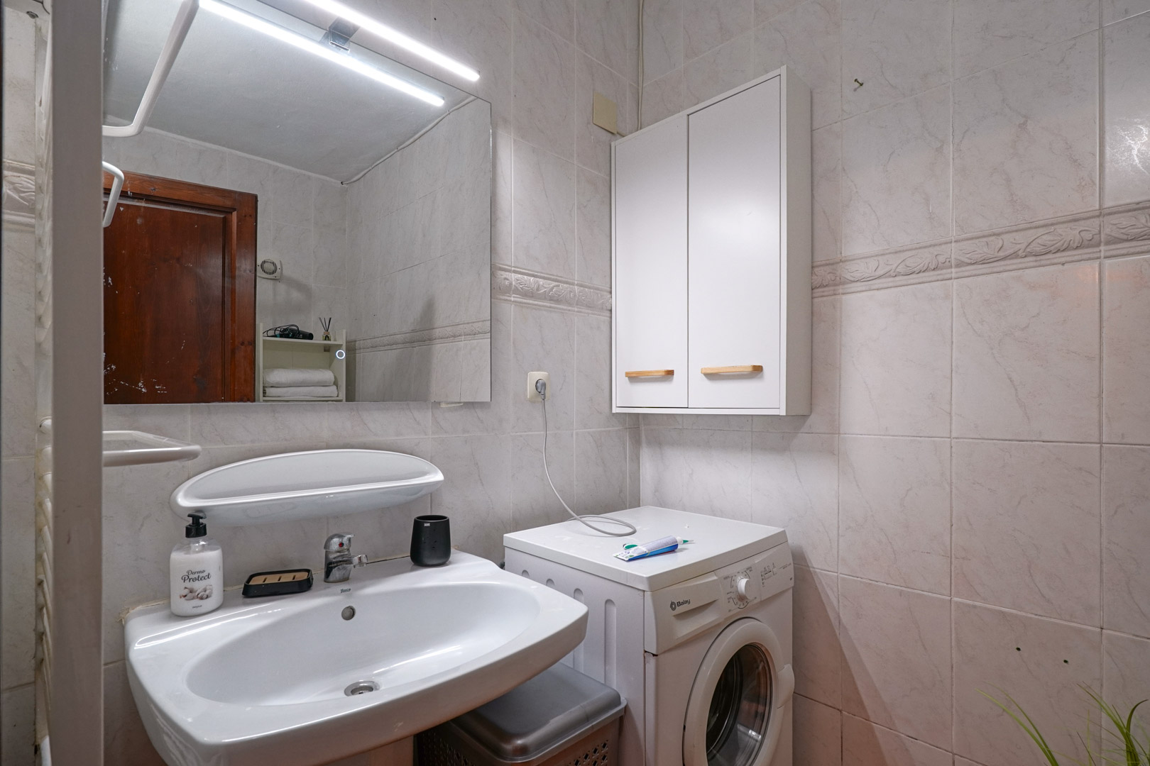 Salle de bain fonctionnelle avec lavabo en céramique, miroir à éclairage LED et coin buanderie intégré comprenant lave-linge et armoire murale de rangement.