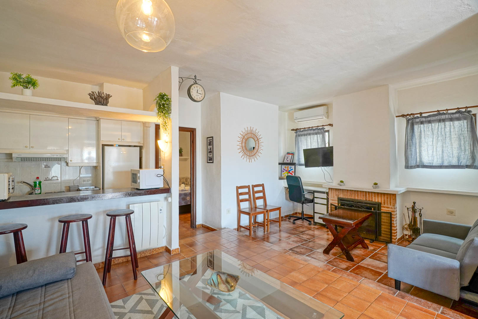 Séjour lumineux avec cheminée en brique, sol en terre cuite et cuisine semi-ouverte équipée d'un bar et de tabourets en bois.
